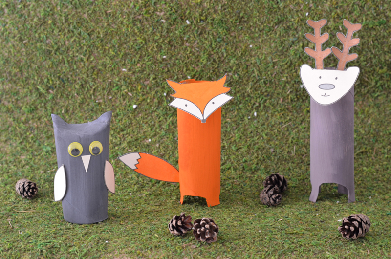 Trois animaux en carton en forme de renne, renard et hibou entourés de pommes de pin dans un décor herbe.