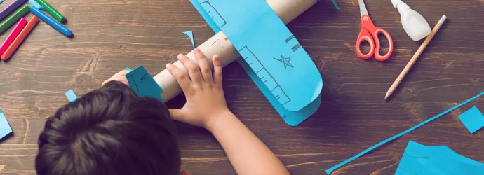 Un enfant qui fabrique un avion en carton avec des ailes et une gouverne de direction bleues, décorées de dessins.