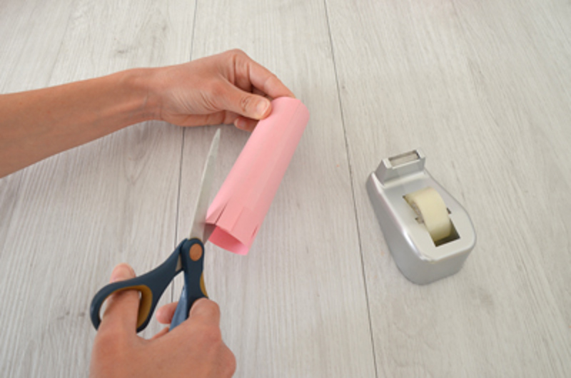 Une personne fait une entaille dans un tube rose couvert de papier coloré avec des ciseaux.