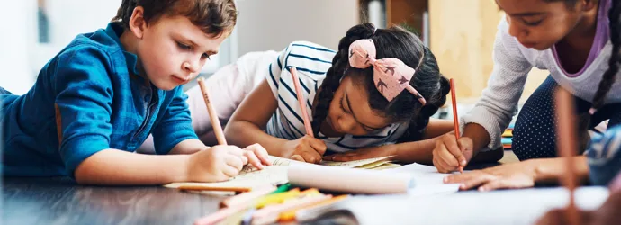 Trois enfants allongés sur le sol dans une maison dessinent dans des livrets avec des crayons.