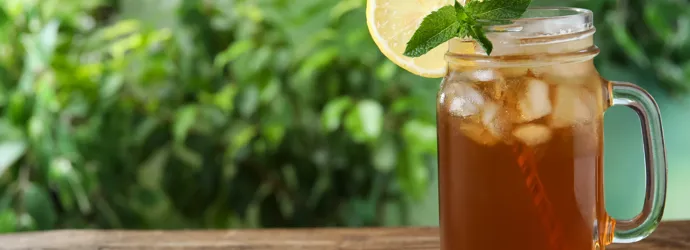Mug en verre transparent posé sur une table en bois dans un jardin, dans lequel se trouve du thé glacé à la menthe. Il y a une tranche de citron sur le bord du mug, et des plantes en arrière-plan.