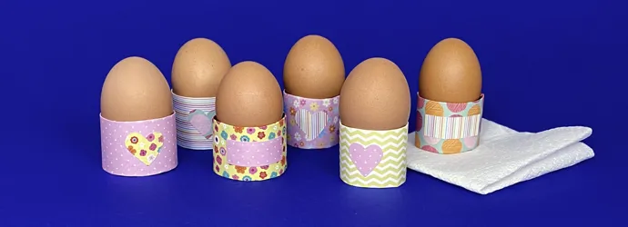 6 oeufs dans des coquetiers de motifs colorés faits maison, sur un fond bleu. Un des oeufs est posé sur un essuie-tout.