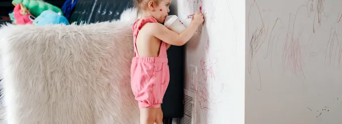 Petite fille habillée en rose dans un salon. Elle dessine au feutre sur le mur d'une main, et boit du lait de son biberon de l'autre main.