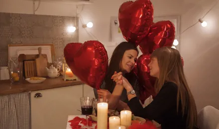 Deux femmes dînant aux chandelles se tenant la main. Il y a des ballons rouges en forme de coeur et des bougies